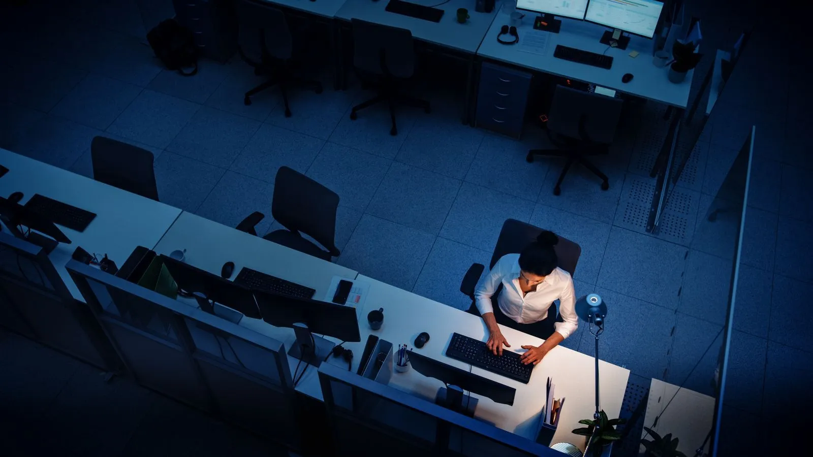 Woman working alone at a desk in a dimly lit office late at night typing on a keyboard.
