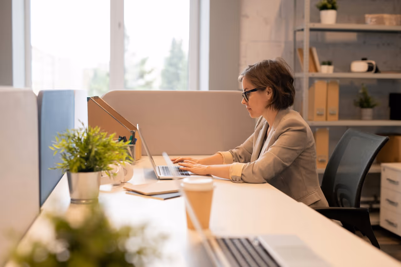 Woman wearing glasses typing on a laptop at a bright office desk with plants and notebooks.
