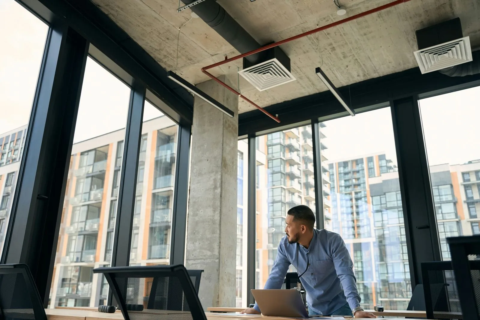 Man in blue shirt leaning on a desk with laptop in a modern office with large windows.