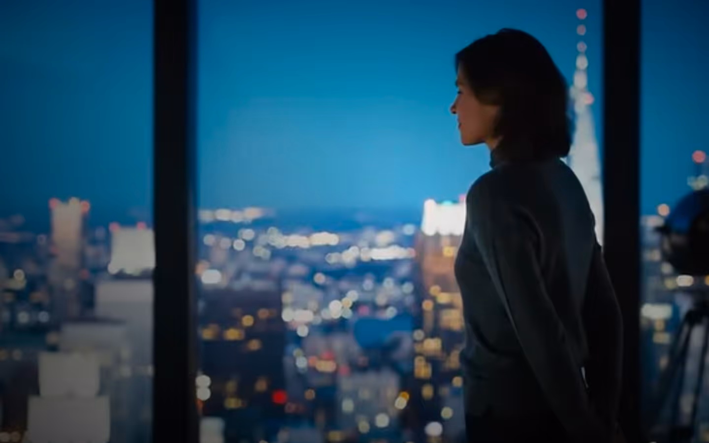 Woman with dark hair in a turtleneck looking out a window at a cityscape at dusk with lights.